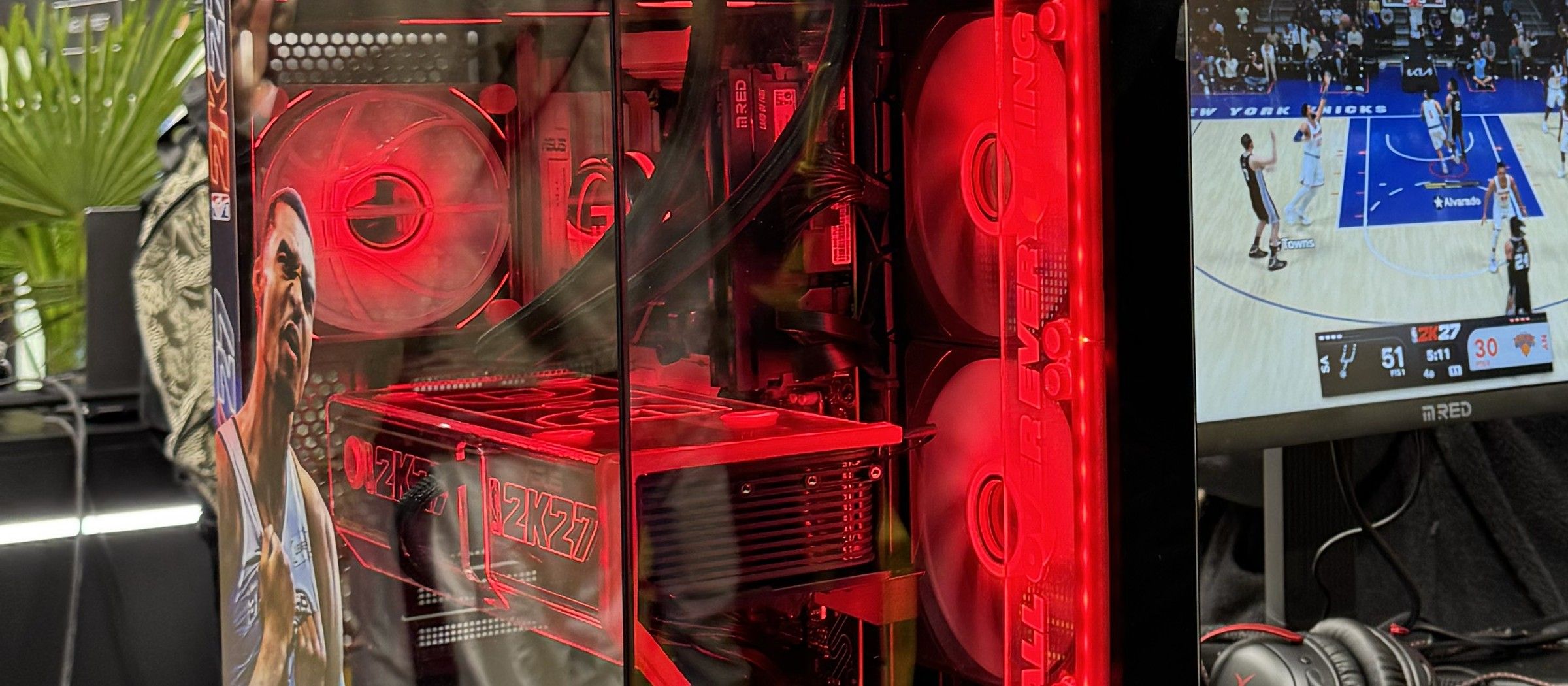 Le flanc vitré du boîtier NBA 2K27, ventilateurs et gravures éclairés en rouge, et à droite l’écran M.RED où se joue une partie